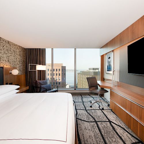 A modern hotel room features a bed, a wall-mounted TV, a desk with a chair, and a large window with a city view in the background.