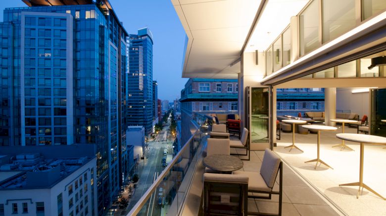 A modern balcony view of a cityscape with tall buildings at dusk. The balcony has tables, chairs, and is illuminated by both natural and artificial light.