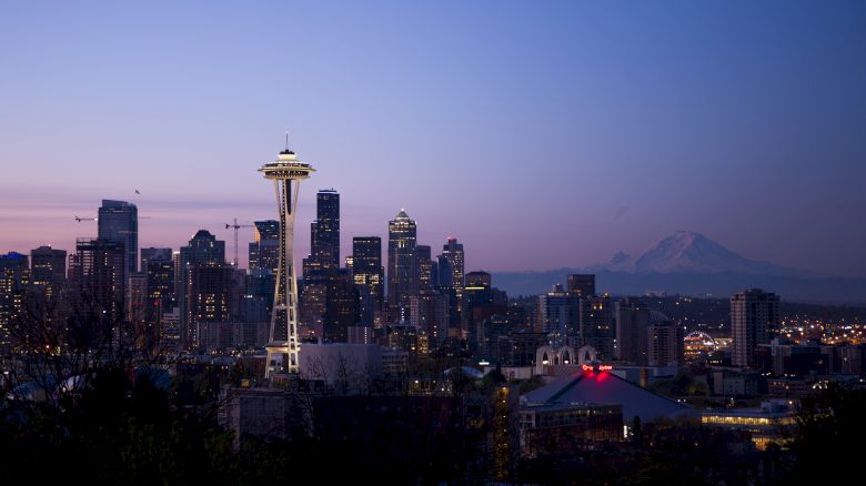 A city skyline at dusk featuring a prominent tower, with mountains visible in the background. The sky is a gradient of blue and purple hues.
