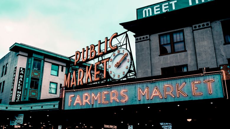 The image shows a well-known farmers market with neon signs reading "PUBLIC MARKET" and "FARMERS MARKET" against an urban backdrop.