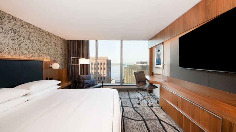 A modern hotel room with a bed, a desk with a chair, a large TV, and floor-to-ceiling windows offering a city view.