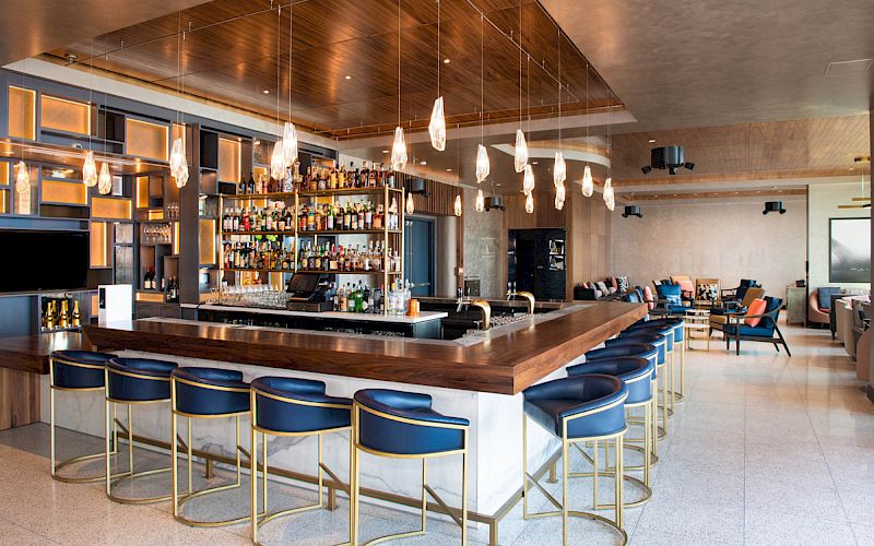 Modern bar with blue barstools, a well-stocked liquor shelf, pendant lights, and a seating area in the background.