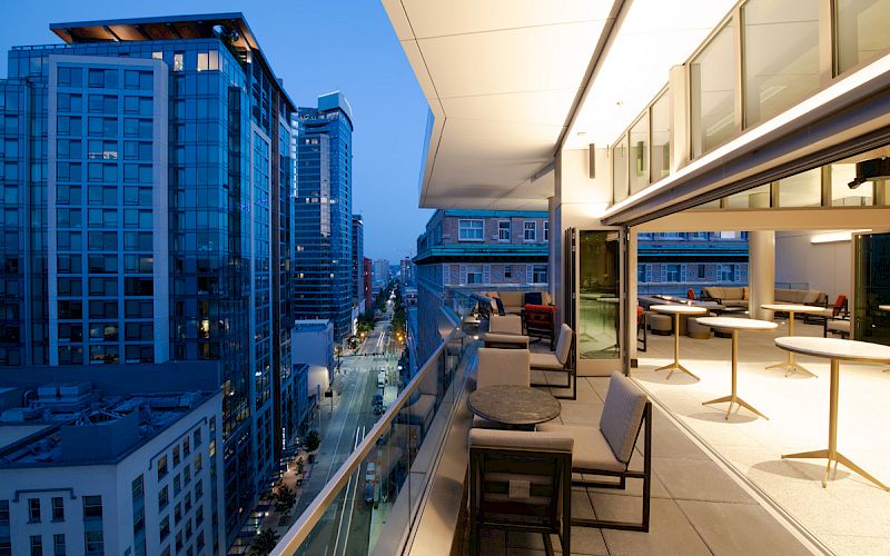 A modern terrace with seating and tables overlooks a cityscape with tall buildings against a twilight sky.
