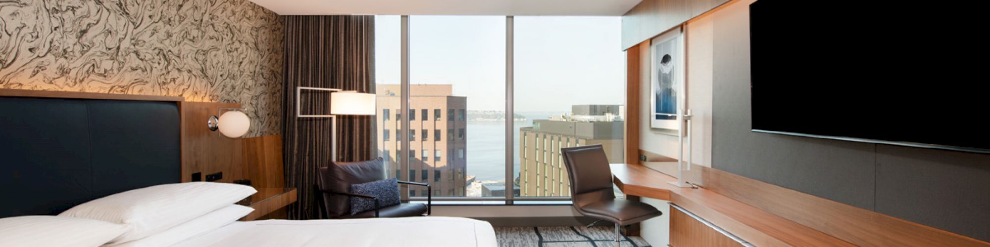 A modern hotel room with a large bed, wall-mounted TV, desk, chair, and a window providing a city and waterfront view.