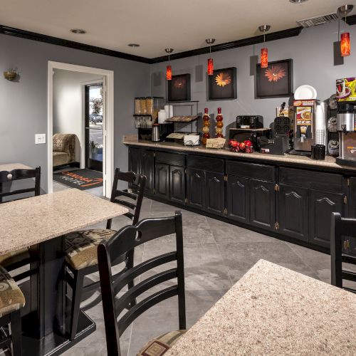 The image shows a small dining area with tables and chairs, coffee machines, and wall decor. It appears to be a cozy café or coffee station.