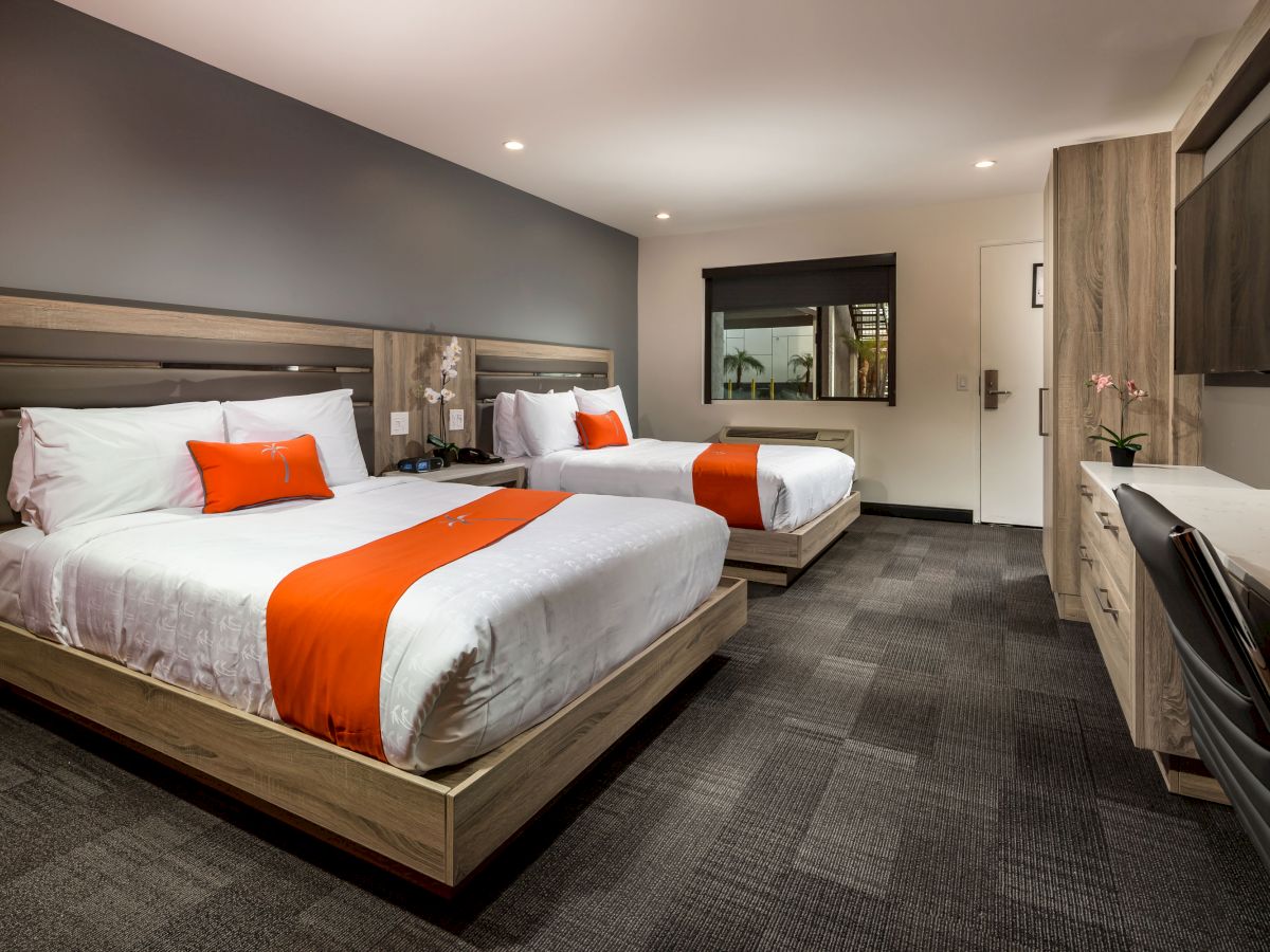 A modern hotel room with two double beds, orange accents, a flat-screen TV, a desk and chair, grey walls, and carpeted flooring.