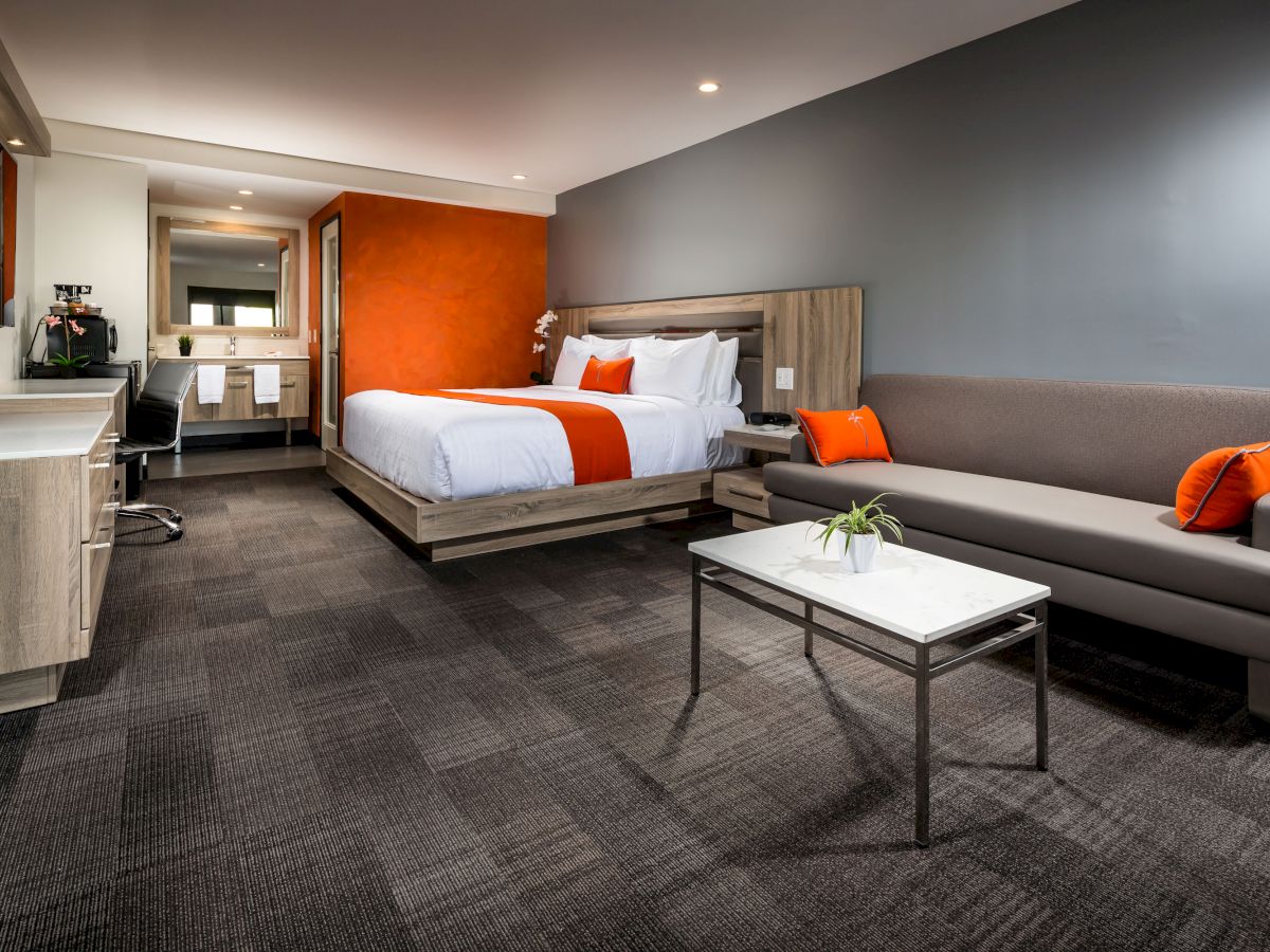 The image shows a modern hotel room with a bed, a couch, a desk, a coffee maker, and a bathroom in the background, all in neutral tones with red accents.