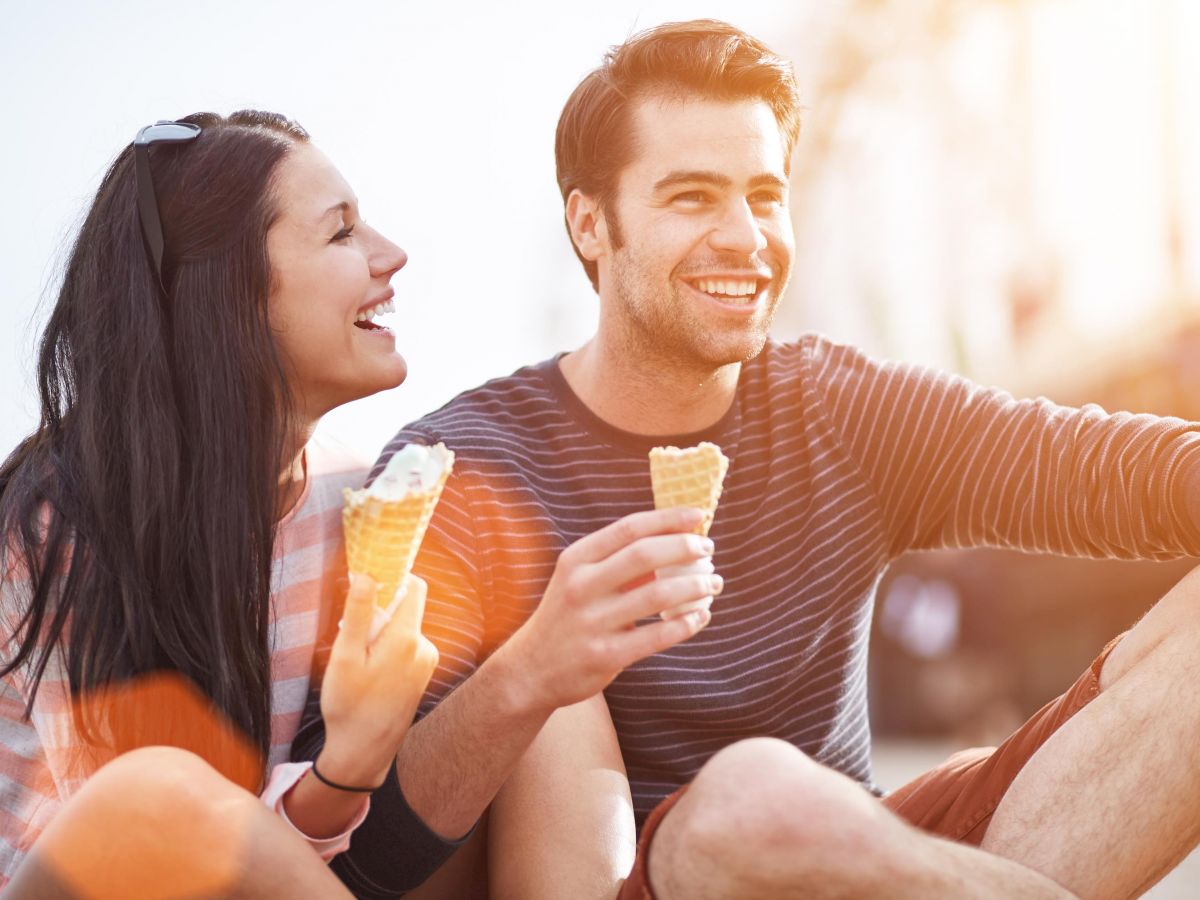 Two people are sitting outdoors, both smiling while holding ice cream cones, enjoying a sunny day.