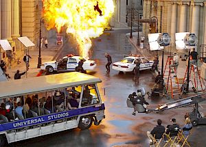 A film set with a Universal Studios tram, police cars, a fire explosion, and production crew equipment, capturing an action scene.