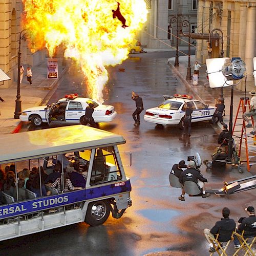 A film set with a Universal Studios tram, police cars, a fire explosion, and production crew equipment, capturing an action scene.