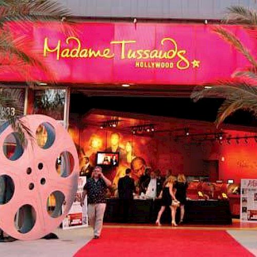 The image shows the entrance to Madame Tussauds Hollywood, adorned with film reel decorations and a red carpet, flanked by palm trees.